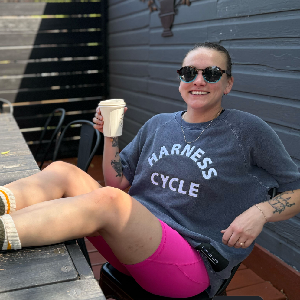 Person sitting outdoors holding a coffee cup, wearing a 'Harness Cycle' shirt.